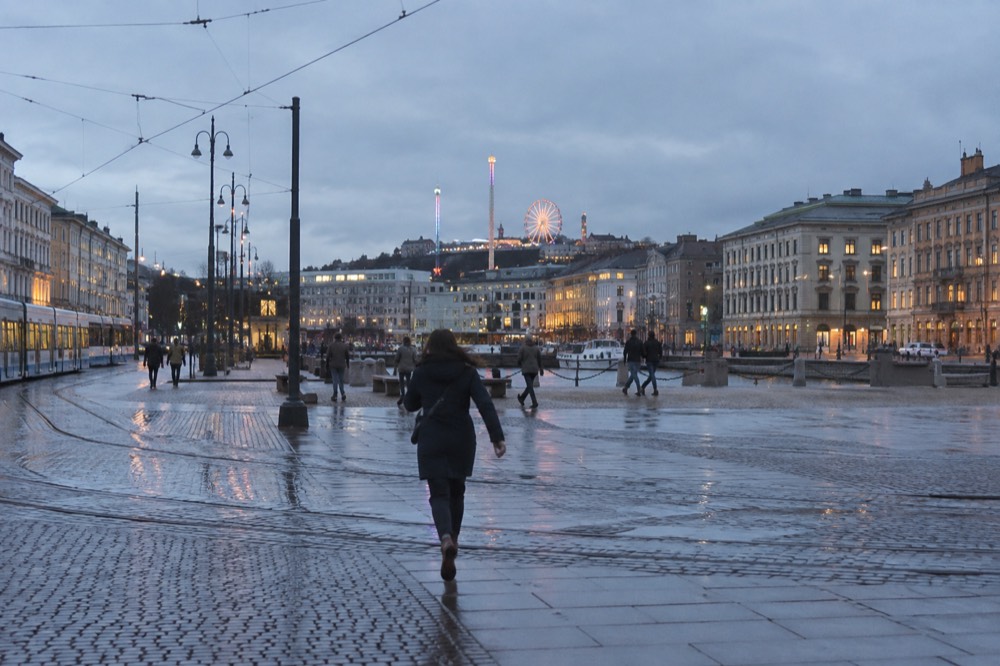Amusement park rides and a Ferris wheel lit up at dusk above a Nordic city skyline