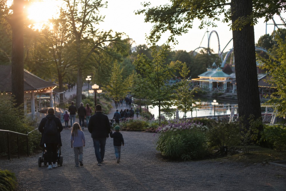 Families walking through a lit amusement park promenade with ride lights in the background