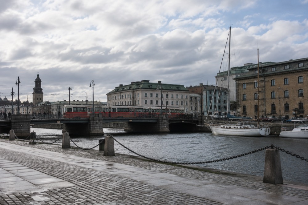 Red trams crossing a stone bridge over a Gothenburg canal, with moored sailboats and brick warehouses behind them