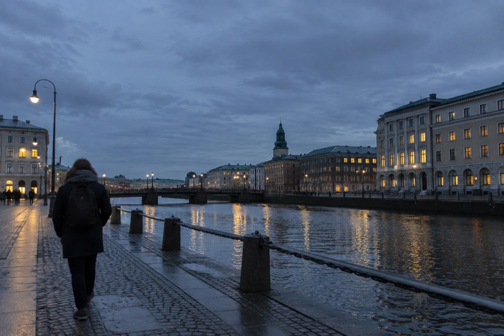 A person in a rain jacket and scarf walking along a Gothenburg canal under a soft grey sky