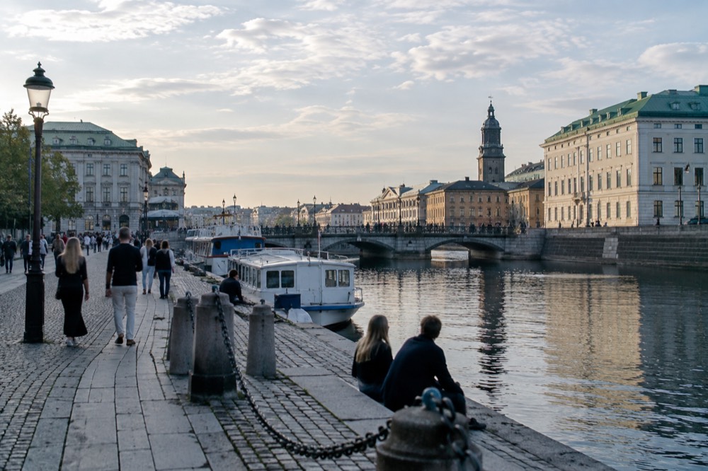 People strolling along a Gothenburg canal on a bright evening with a bridge and moored boats behind them