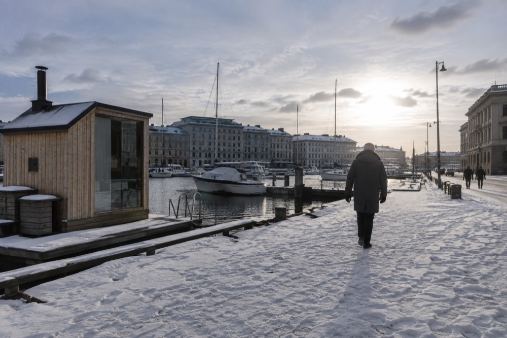 A wooden sauna hut beside a snowy harbour with an ice-free channel and small boats