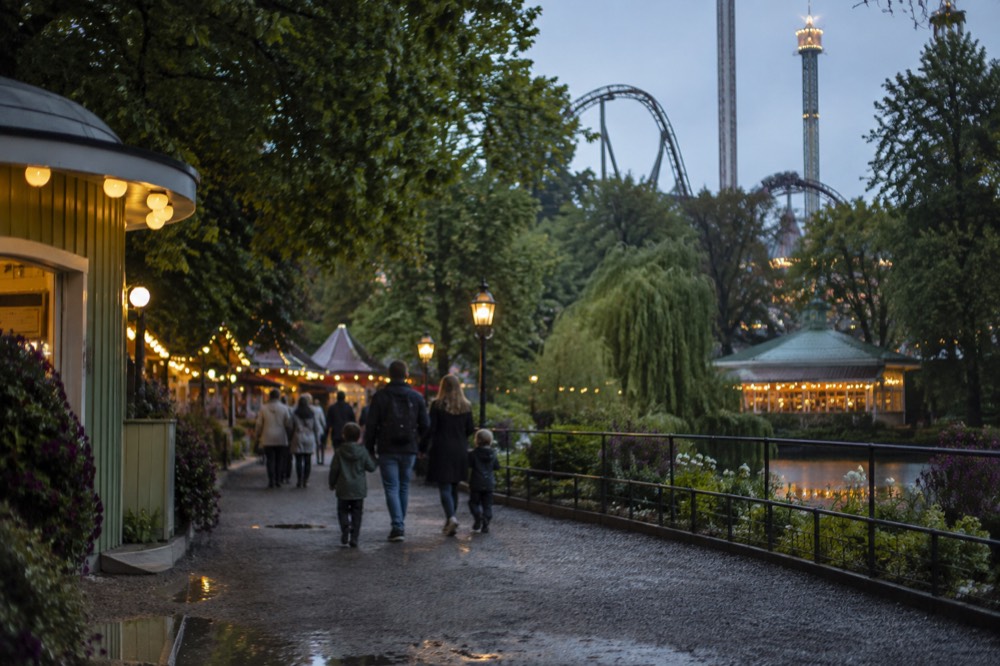 Families walking down a lit pathway between amusement rides and food stalls in an amusement park at dusk
