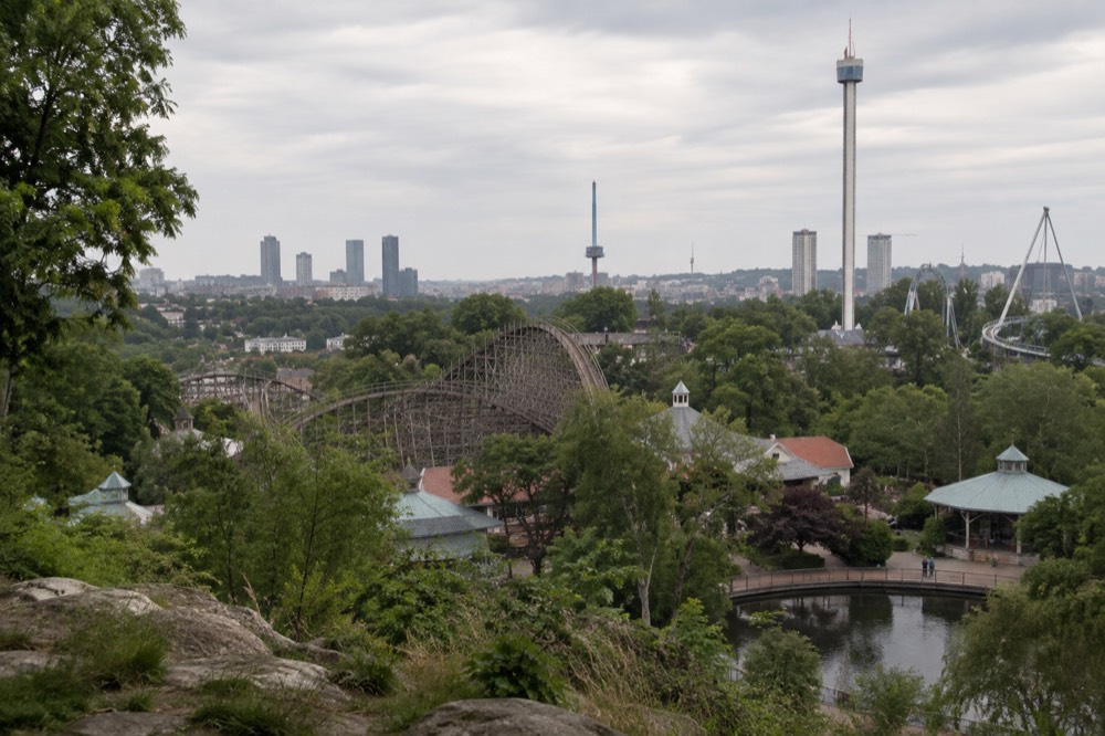 A large amusement park seen from above, with a wooden roller coaster curving between tree-lined paths and the towers of Gothenburg beyond