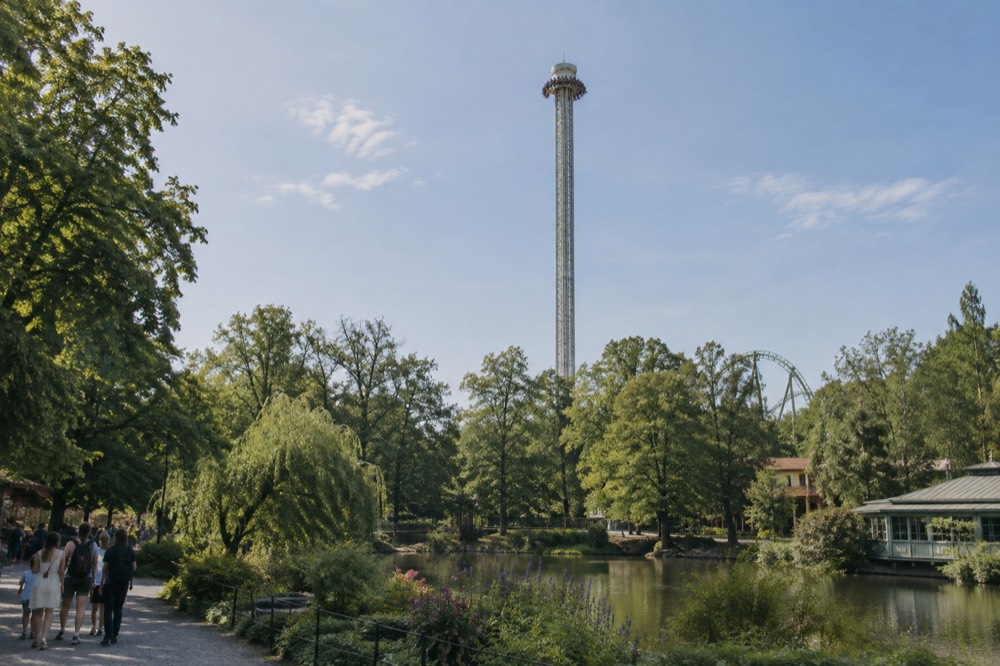 A tall drop tower ride seen from below, with riders at the top of the tower against a blue sky