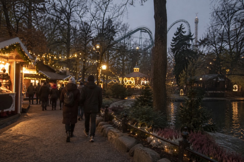 A park pathway at night hung with strings of warm lights, with people in coats walking between lit market stalls