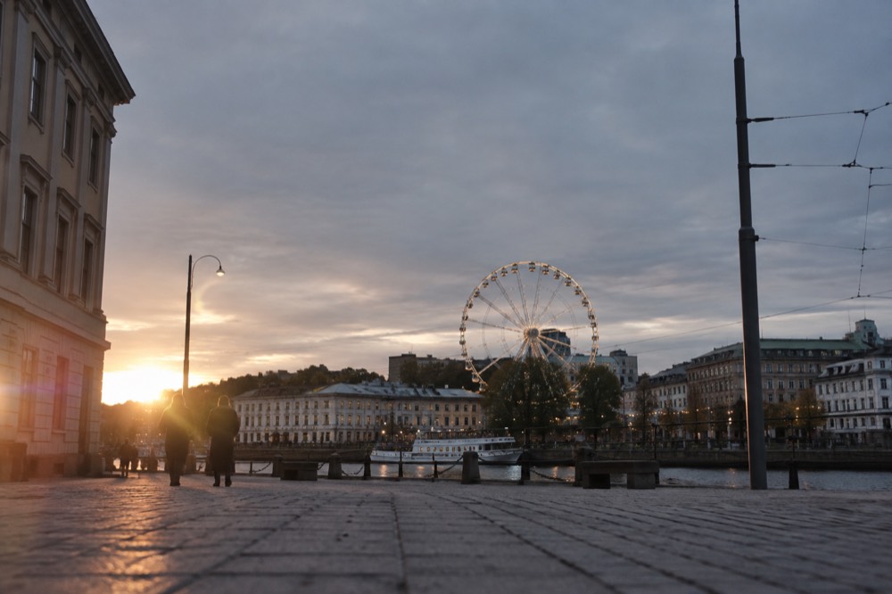 A ferris wheel with lit gondolas turning above a city skyline at dusk, with apartment buildings and a river below
