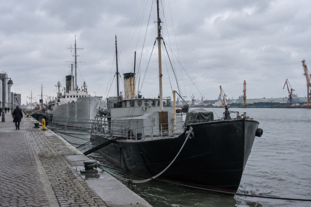 Historic ships moored together on a city quay, masts and funnels against a grey Nordic sky