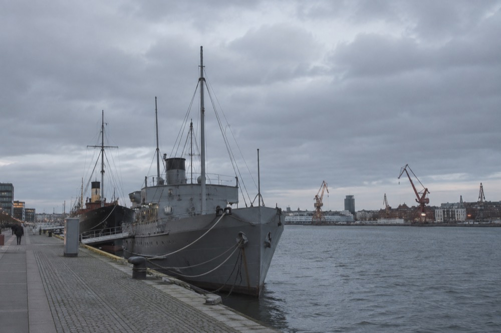 Museum ships moored at a quay in warm evening light with the city skyline behind