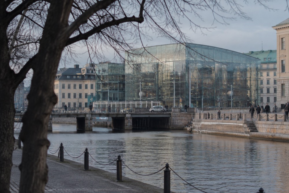 A glass science centre building seen from a canal path with trees in the foreground