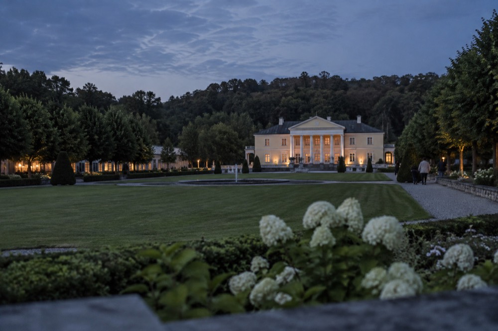 A white neoclassical country house with a columned portico seen across a lawn, with tree-lined gardens on either side