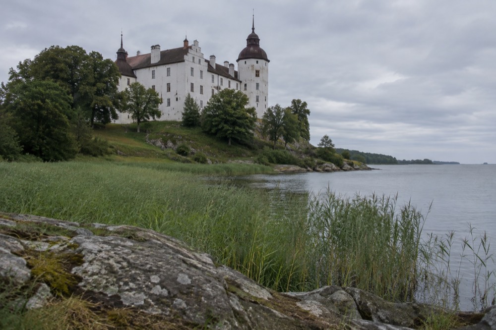 A stone castle with a tall round tower rising from a green headland above a wide open lake