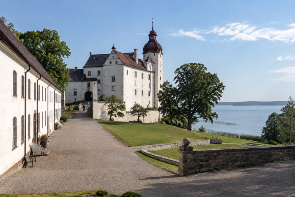 A long stone castle courtyard with arched windows and green lawns leading down to the lake