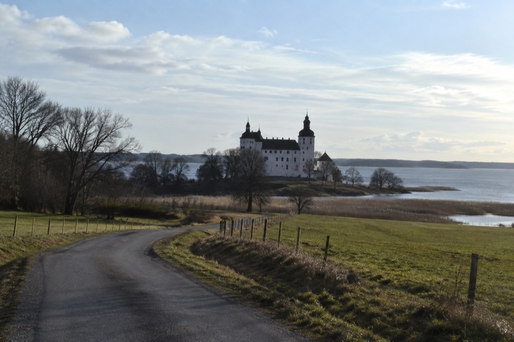 A country road curving through green farmland toward distant water under a bright sky