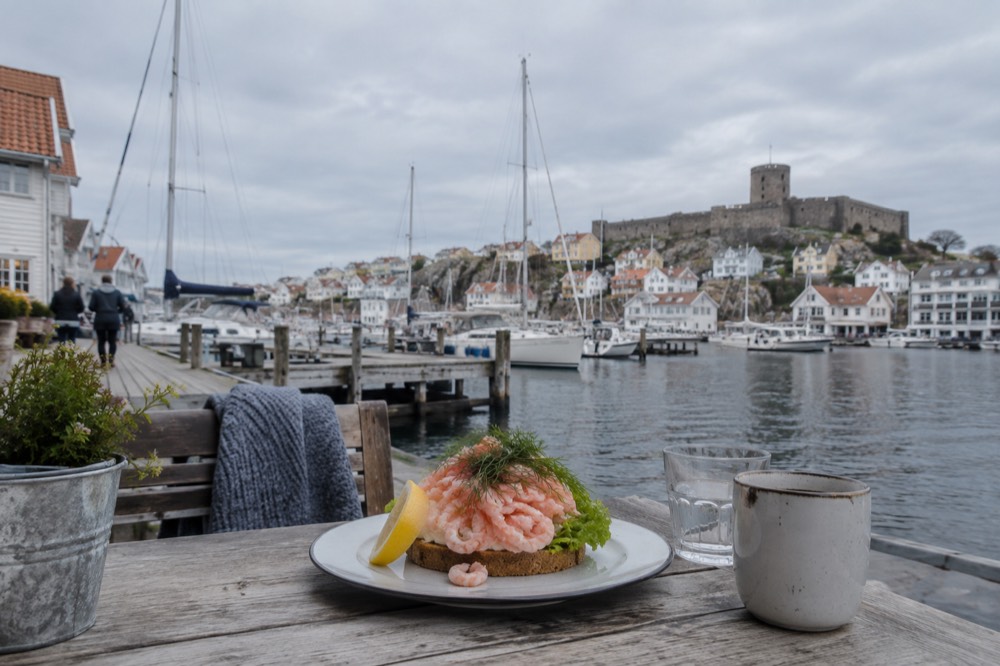 A plate of peeled shrimp on bread at an outdoor harbour table with boats behind