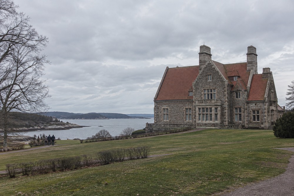 A stone manor house with a steep red-tiled roof seen across a lawn sloping toward a narrow sea inlet