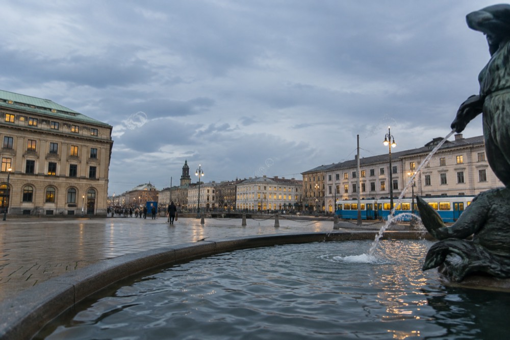 A city square with fountains and stone buildings in evening light