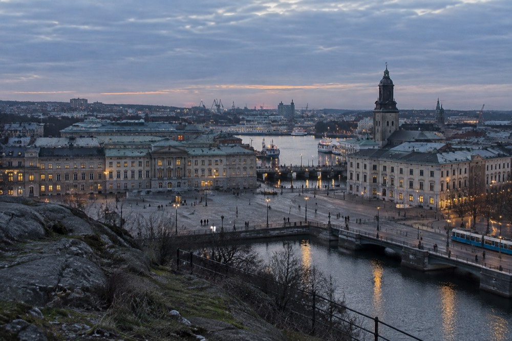 A panoramic city view from a rocky hilltop at dusk, with a river and church towers below