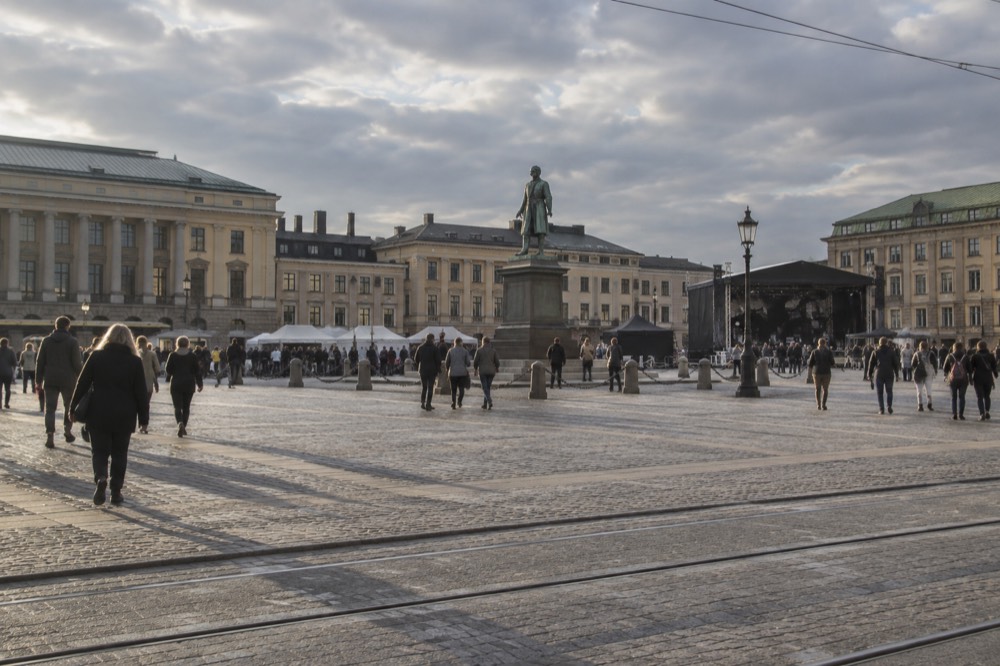 A wide plaza with a bronze statue at its centre, a concert hall on one side and crowds crossing between festival stages