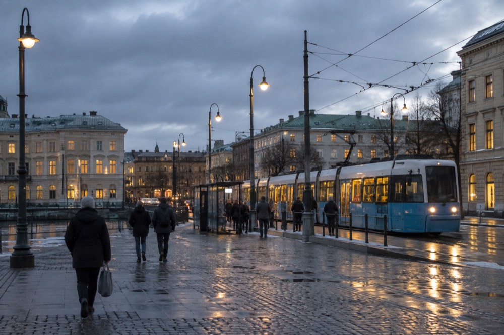 A Gothenburg tram stop at dusk with lit tram and wet pavement reflecting the lights