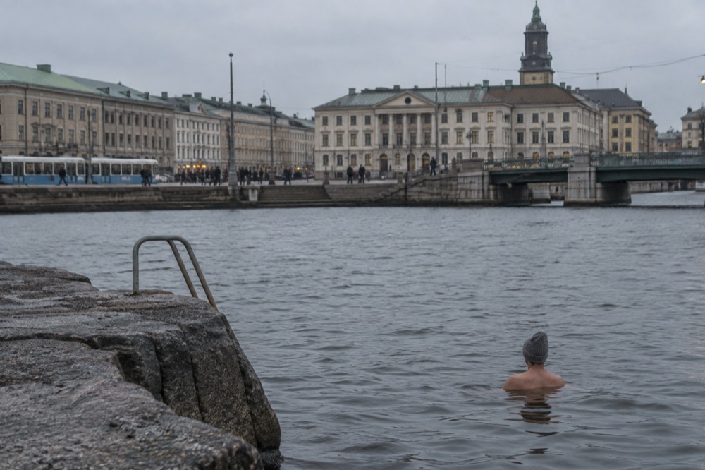 A swimmer in a wool hat standing in calm grey sea beside a granite rock ledge
