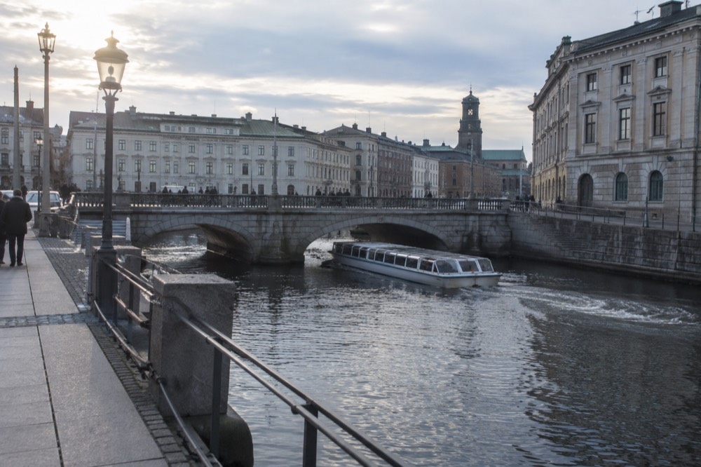 A sightseeing boat passing under a low stone bridge on the Gothenburg canal with old buildings on both banks