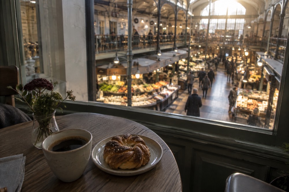 A cup of black coffee and a cardamom bun on a small wooden cafe table beside a window
