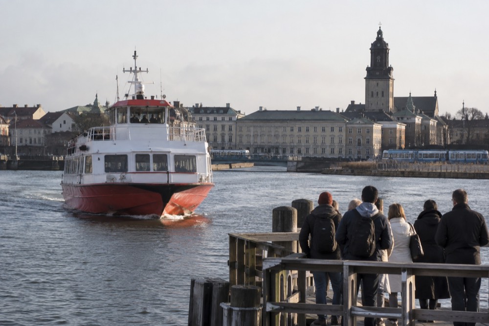A red-and-white archipelago ferry approaching a wooden jetty on a green island, passengers waiting