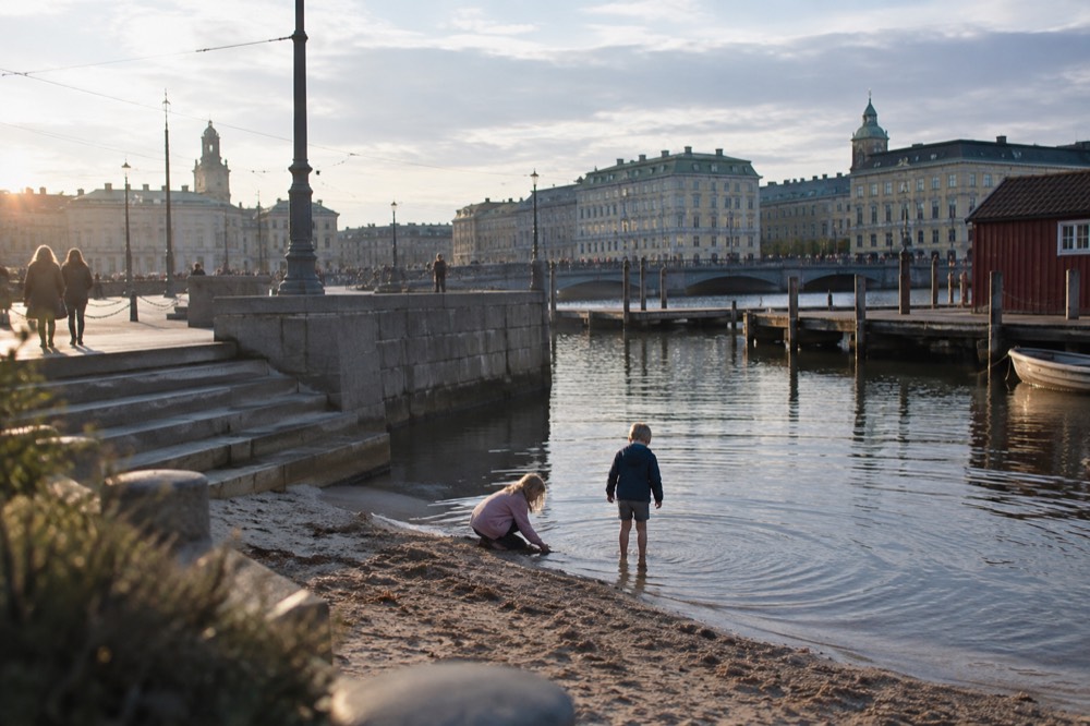 Children playing in shallow clear water at a small sandy bay, wooden jetty and red boathouse behind