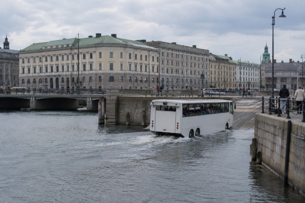 Amphibious tour bus driving down a ramp toward harbour water beside a stone quay